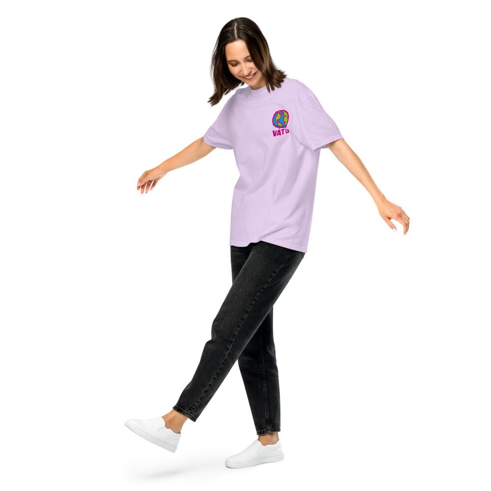 A woman in a light purple tee with a colorful globe and "VATO" design, black jeans, and white shoes, balancing on one foot.